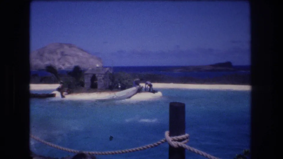 1977 Honolulu, Oahu 8mm frame of a marine show lagoon after a jump, with distant people on the sandy set, a small structure, plants, and rope barrier in the foreground.