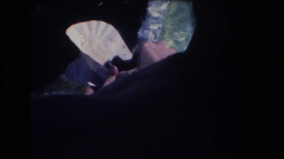 1977 Honolulu 8mm frame showing a person's hand holding a pale folded item or food near dark shoreline edges above blue-green water.