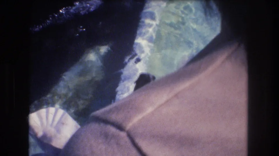Downward 1977 home-movie view of blue-green water with a pale object near the lower edge and part of a person's tan garment in the foreground.