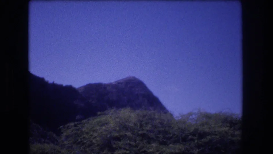1977 Honolulu home-movie frame showing a low mountain ridge, dense green trees, and a clear blue sky in a tropical landscape view.