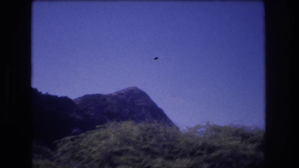 1977 Honolulu home-movie frame with a low mountain ridge and leafy treetops beneath an open blue sky, with a small dark film speck visible in the sky area.
