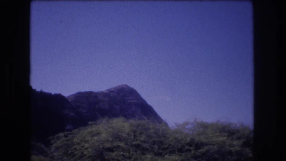 Handheld 8mm view from Honolulu, Oahu in 1977 showing a dark mountain peak, foreground brush, and blue sky within rounded film-frame edges.