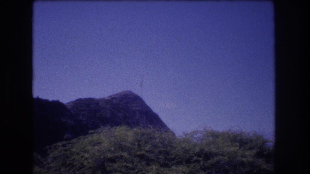 1977 Oahu travel-film frame with a low dark ridge, green vegetation, blue sky, and visible 8mm grain and color shift.
