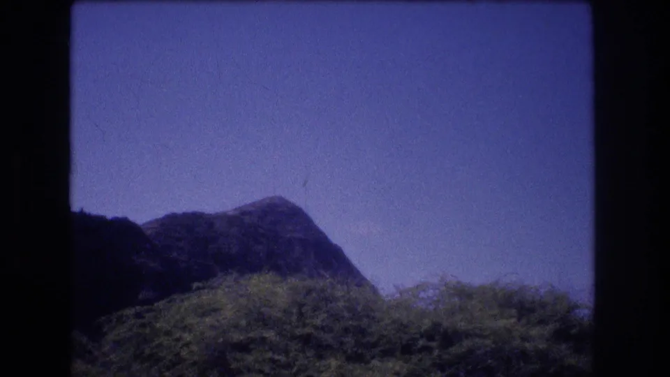 1977 Oahu travel-film frame with a low dark ridge, green vegetation, blue sky, and visible 8mm grain and color shift.