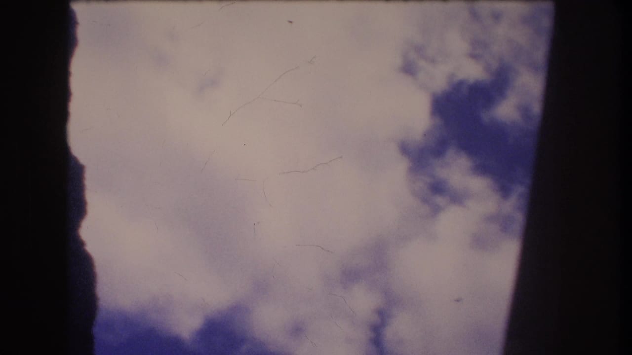 1977 Honolulu 8mm home-movie frame showing blue sky and white clouds viewed through dark vertical edges, with visible film scratches and soft exposure.