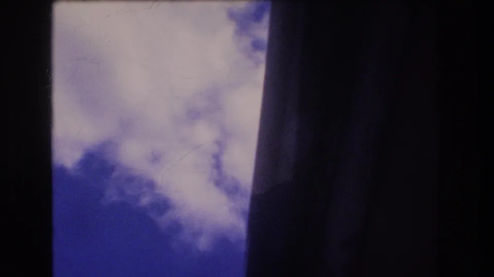 1977 Honolulu 8mm still with white clouds and blue sky visible through a narrow framed opening, bordered by dark vertical shapes and light film wear.