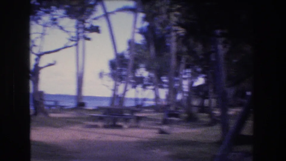 Handheld 1977 Oahu home-movie frame showing empty outdoor tables in a tree-lined clearing, with the ocean visible through soft focus and color shift.