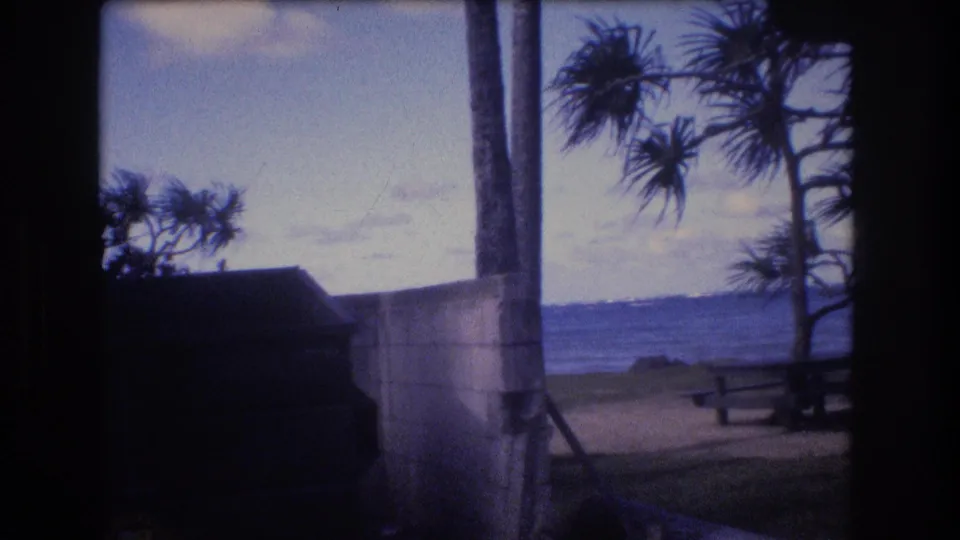 1977 Honolulu-area 8mm frame showing a concrete wall and vertical pole near a beach, with a picnic table, coastal trees, and ocean beyond.