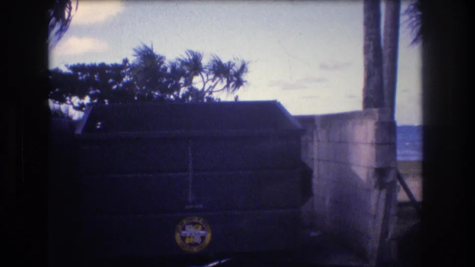 1977 Honolulu-area 8mm frame with a large dark dumpster bearing a circular logo beside a concrete wall, with trees and ocean visible behind it.