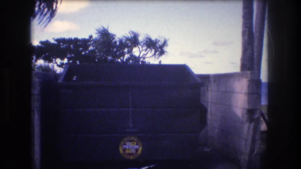 1977 Honolulu-area 8mm frame centered on a dark beachside dumpster with a circular logo, backed by a concrete wall, coastal trees, and the ocean horizon.