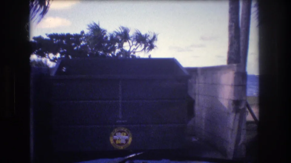 1977 Honolulu Oahu 8mm frame showing a dark dumpster with a circular logo beside a concrete beach wall, with palm foliage and ocean visible in the background.