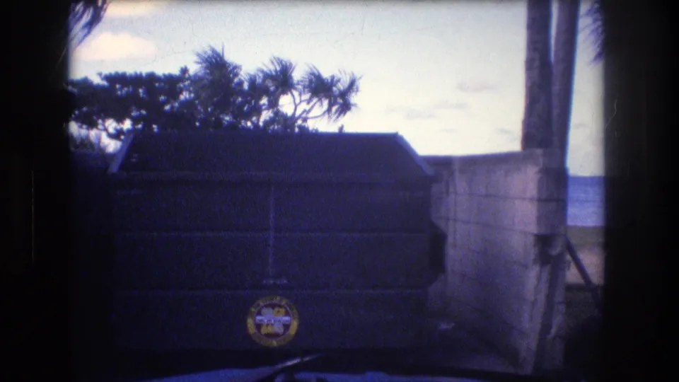 1977 Honolulu Oahu 8mm frame showing a large dark dumpster with a circular logo, a concrete wall, palm foliage, bright sky, and ocean behind the beachside area.