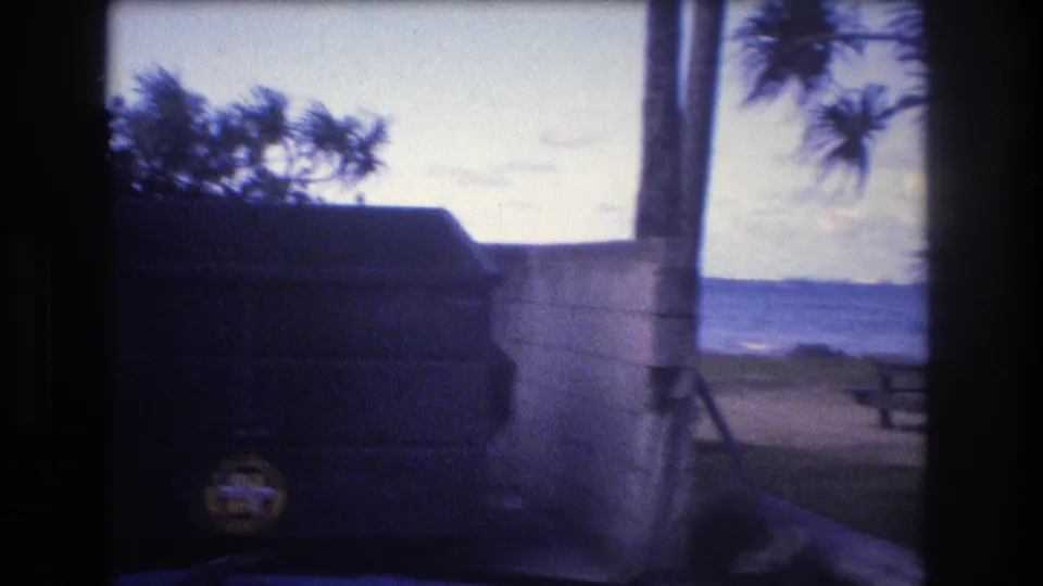 Final 1977 Honolulu Oahu frame with the dumpster partly leaving view beside a concrete wall, revealing palms, ocean, beachside ground, and a small picnic-table form in the distance.