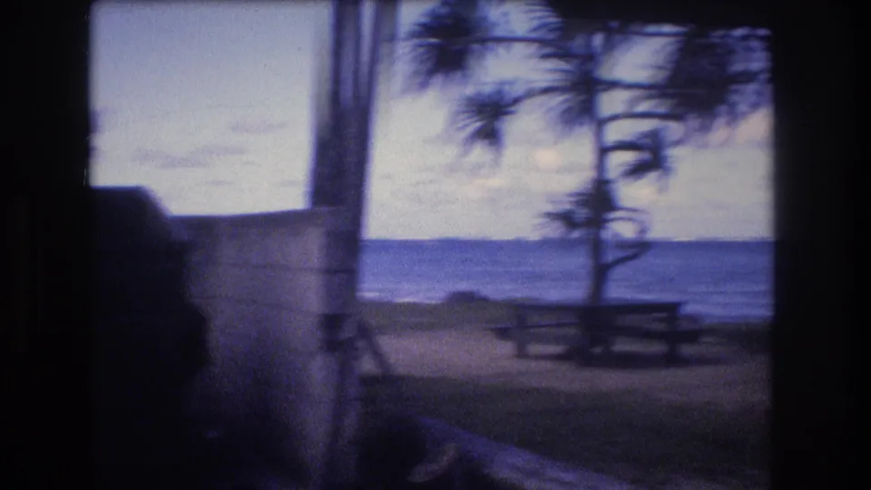 1977 8mm home-movie frame from Oahu showing a shaded campsite view toward a picnic table, wind-shaped coastal trees, and the ocean beyond.