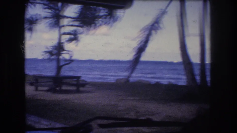 A dark interior or shelter edge frames a 1977 Oahu shoreline campsite with a picnic table, palms or coastal trees, and open water beyond.