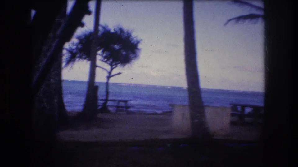 A wider 1977 Oahu campsite view shows coastal trees, picnic tables, light-colored barriers, and the ocean stretching across the background.