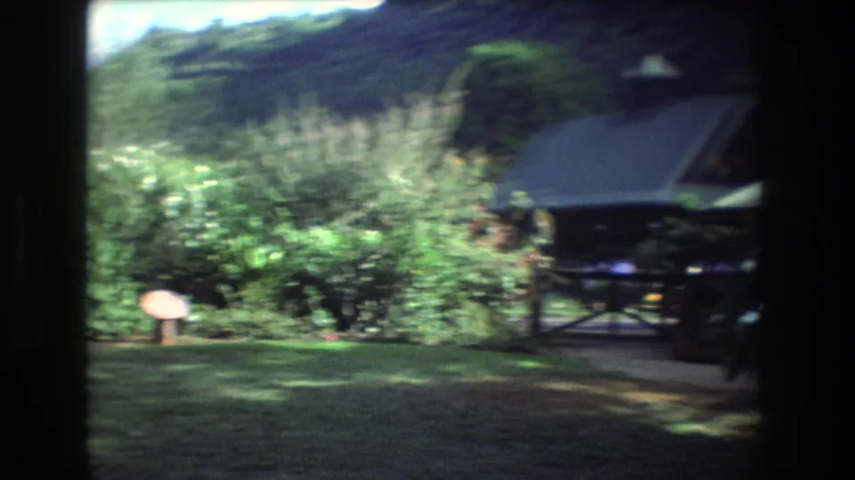 A 1977 Honolulu, Oahu 8mm frame shows a tropical garden area with thick shrubs, green hills, small signs, and a small pavilion or cabin-like structure at right.