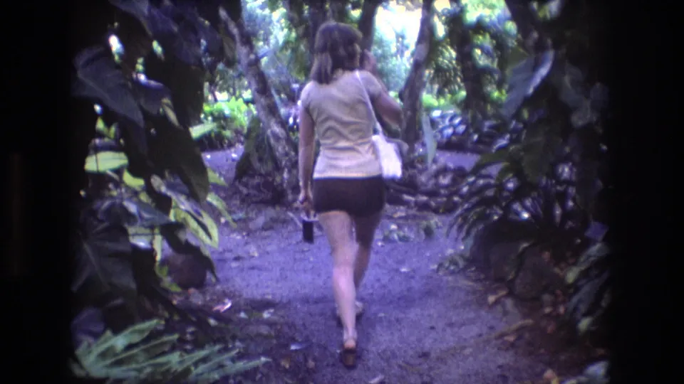 Rear-view 1977 Honolulu home-movie frame following a woman carrying a small bag along a shaded dirt or gravel path surrounded by tropical vegetation.