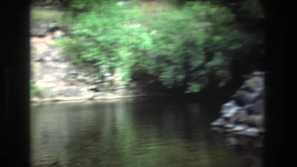 A handheld 8mm view shows lake water, rocky shoreline, and green foliage, with motion blur from the camera movement.