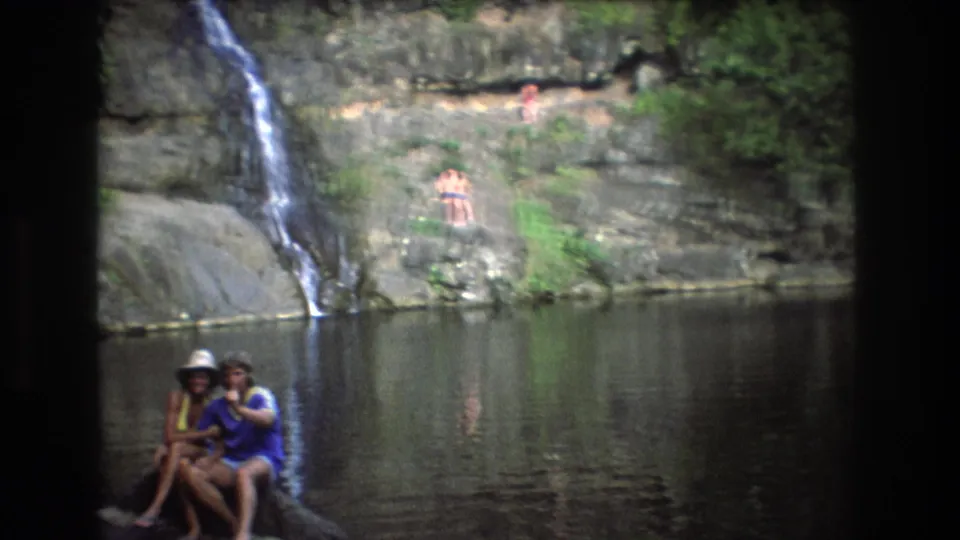 People relax on rocks and sit near the edge of still lake water beneath a rocky ledge and small waterfall in Honolulu, Oahu footage.