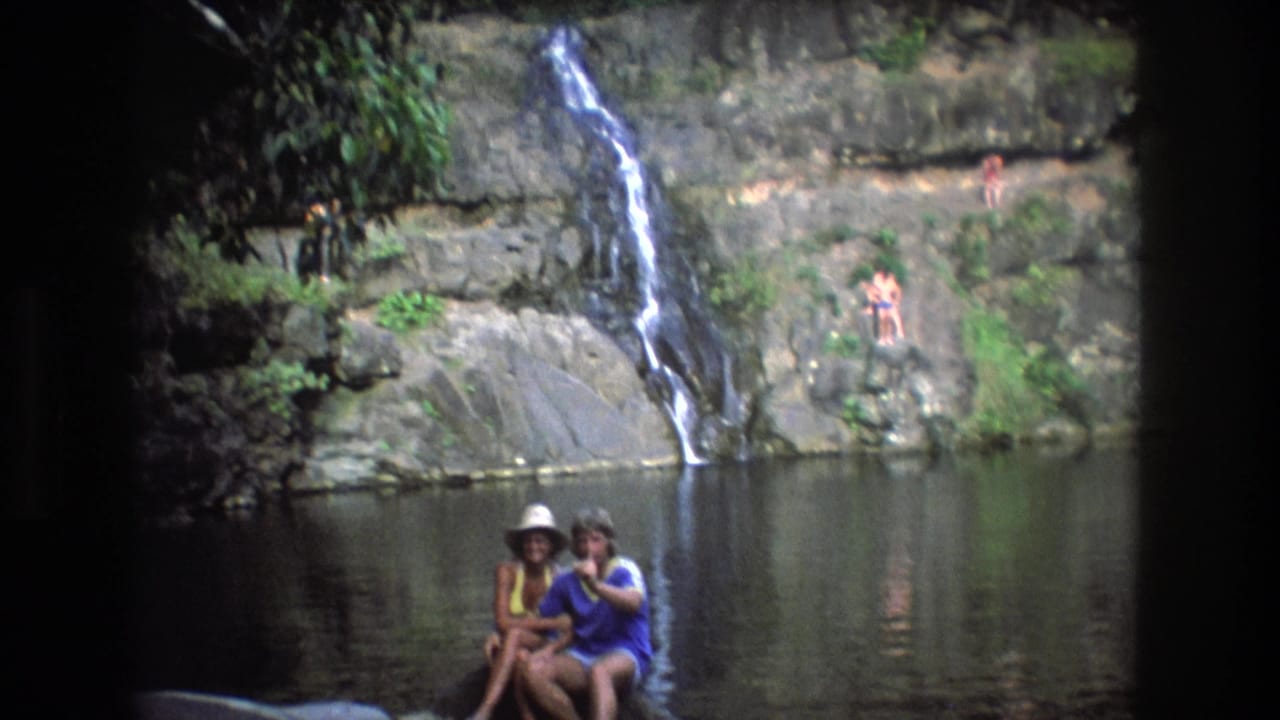 1977 Honolulu, Oahu 8mm home-movie frame showing a small waterfall, rocky ledges, a calm pool, and two adults seated near the water in swimwear.
