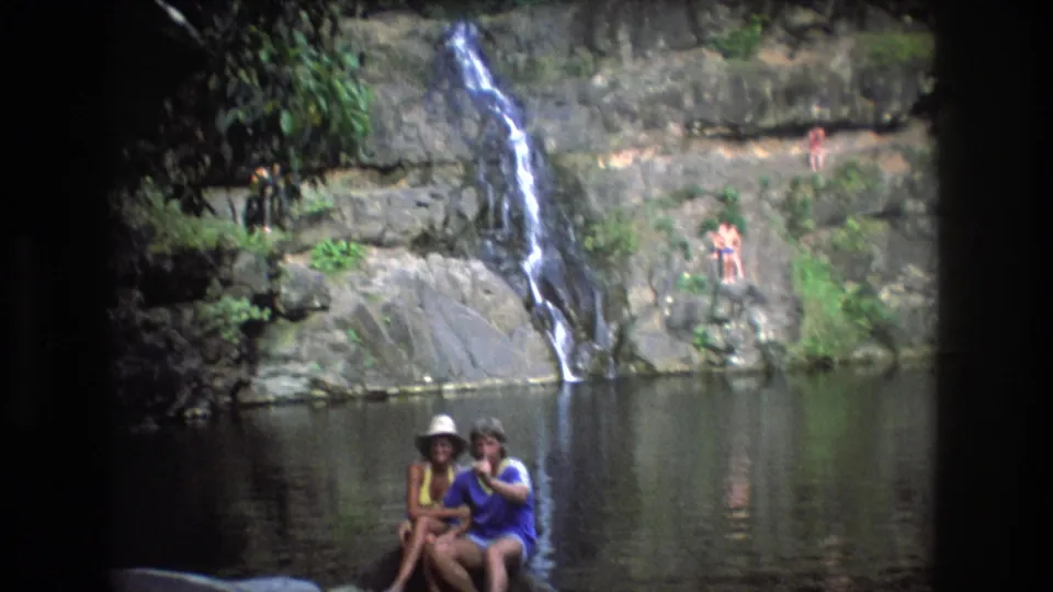 1977 Honolulu, Oahu 8mm home-movie frame showing a small waterfall, rocky ledges, a calm pool, and two adults seated near the water in swimwear.