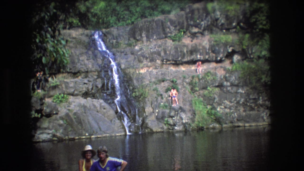 1977 Honolulu, Oahu home-movie frame with two adults in the foreground, distant bathers on rocks, and a waterfall running into a shaded pool.