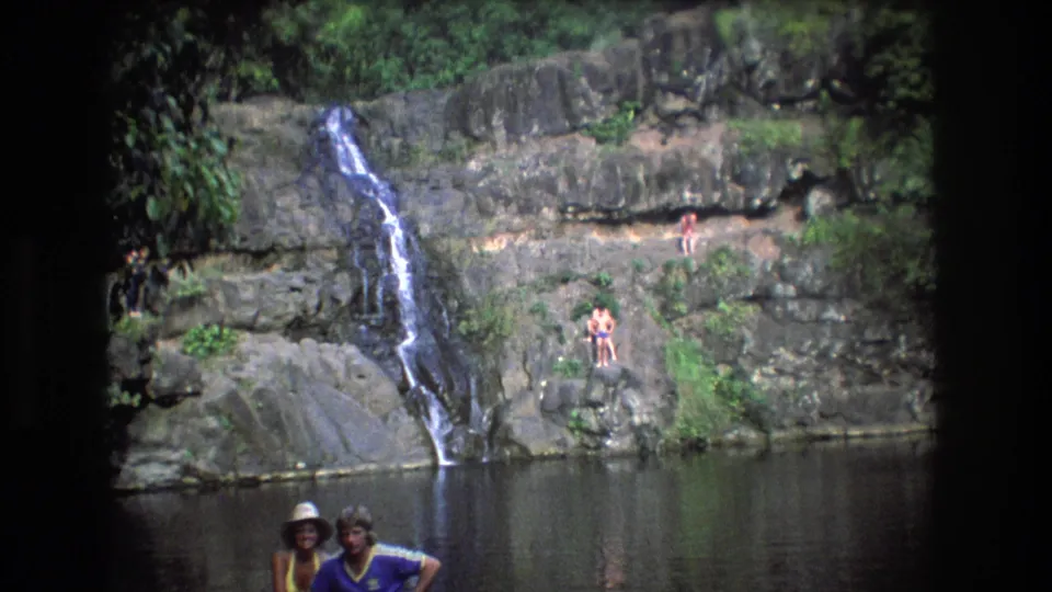 1977 Honolulu, Oahu home-movie frame with two adults in the foreground, distant bathers on rocks, and a waterfall running into a shaded pool.