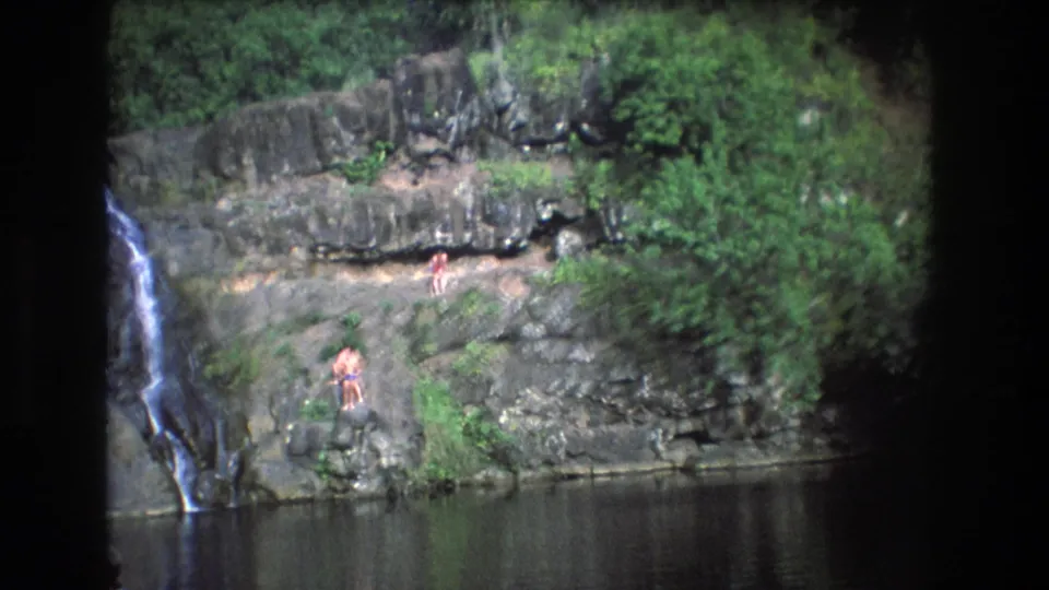 1977 Honolulu, Oahu 8mm frame showing the waterfall at left, a broad rock face, calm water below, and several bathers positioned on the ledges.