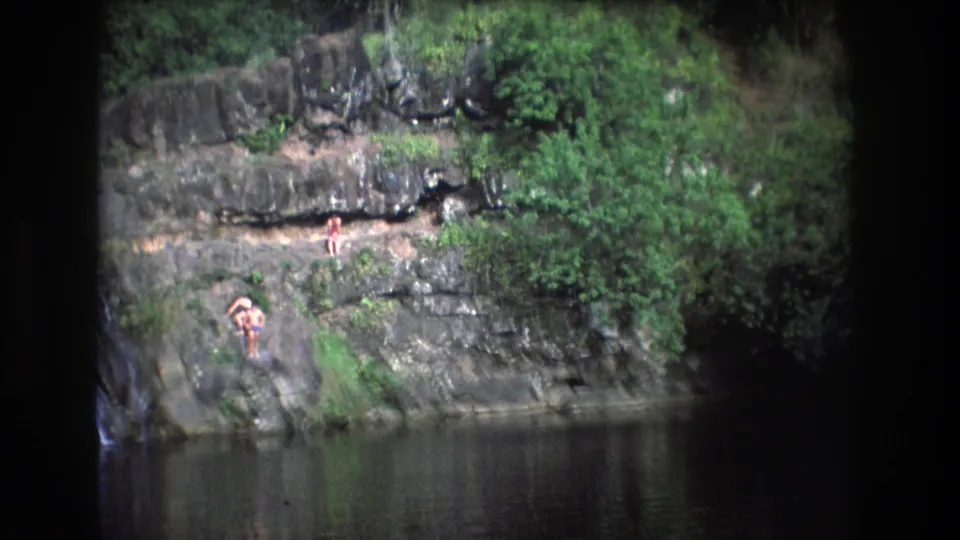 1977 Honolulu, Oahu home-movie frame with distant people on rock ledges, a narrow waterfall at the left edge, a dark pool below, and dense tropical greenery around the scene.
