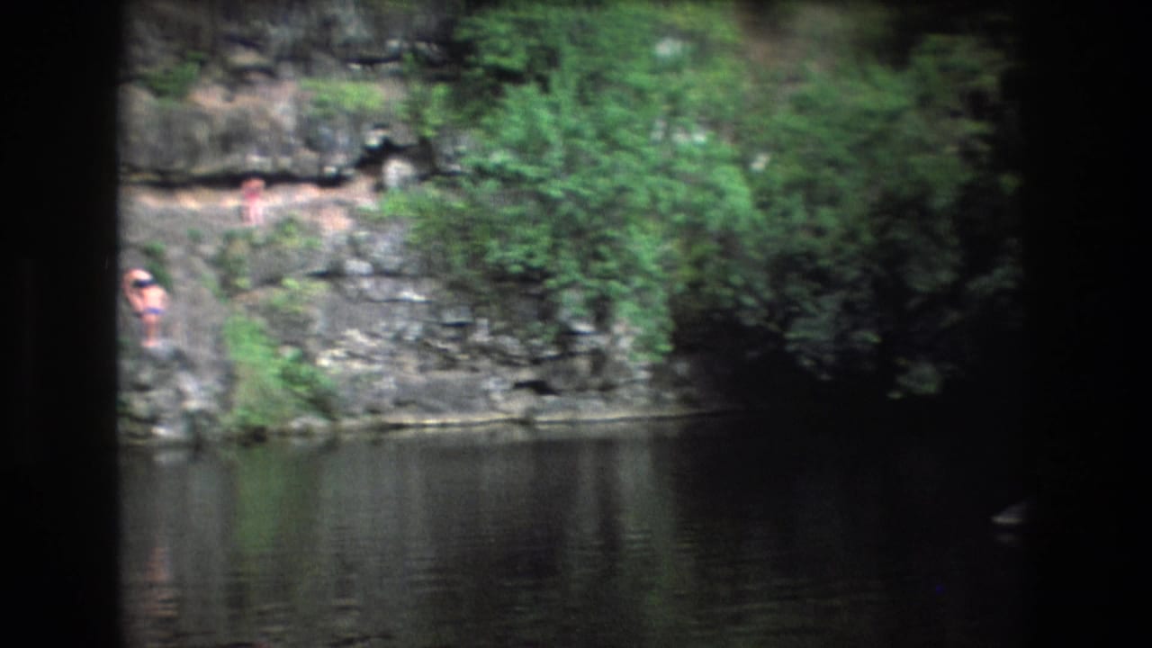 1977 Honolulu, Oahu home-movie frame showing a rocky creek edge, reflective water, dense greenery, and small distant figures near the bank.