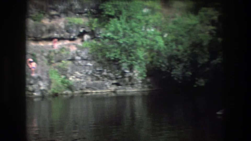 1977 Honolulu, Oahu home-movie frame showing a rocky creek edge, reflective water, dense greenery, and small distant figures near the bank.