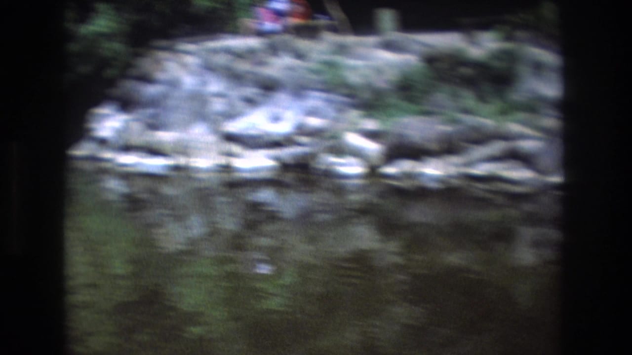 Blurred rocky stream bank with water in the foreground and a small figure near the top edge.