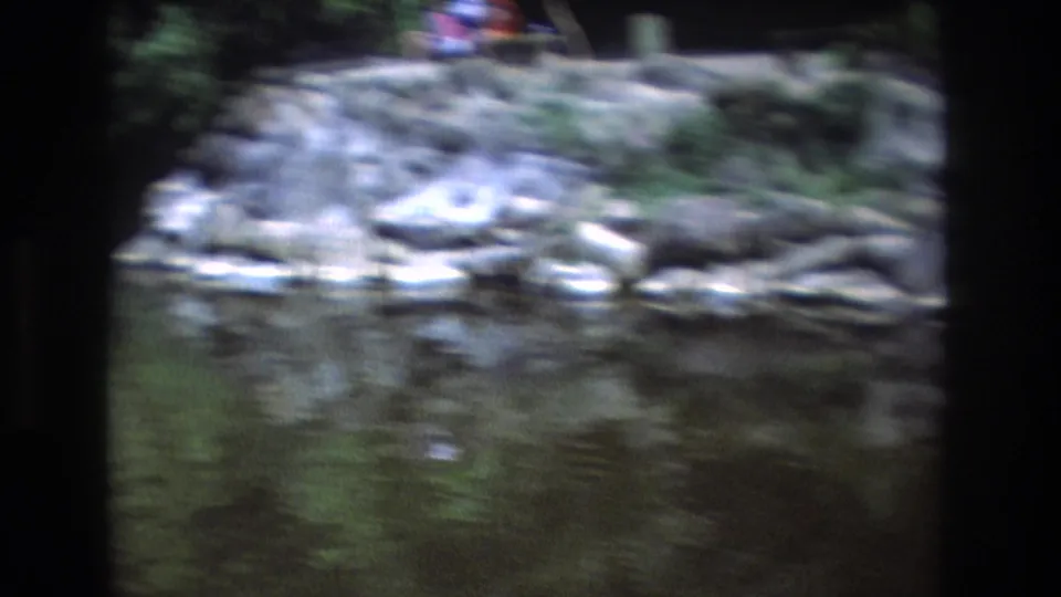 Soft 8mm frame from Honolulu, Oahu in 1977 showing creek water, pale rocks, greenery, and a small distant person near the upper bank.