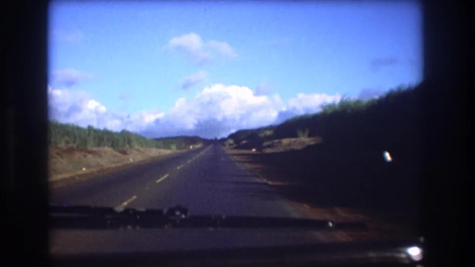 1977 Honolulu, Oahu home-movie frame looking through a car windshield at a paved road, low hills, blue sky, and scattered clouds.