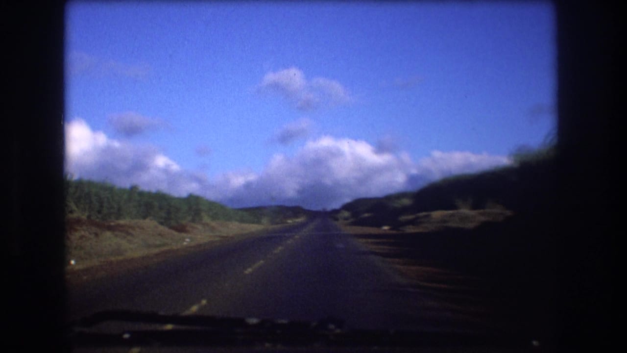1977 Oahu home-movie frame showing a windshield view down a two-lane asphalt road bordered by green vegetation under a bright blue, cloud-filled sky.