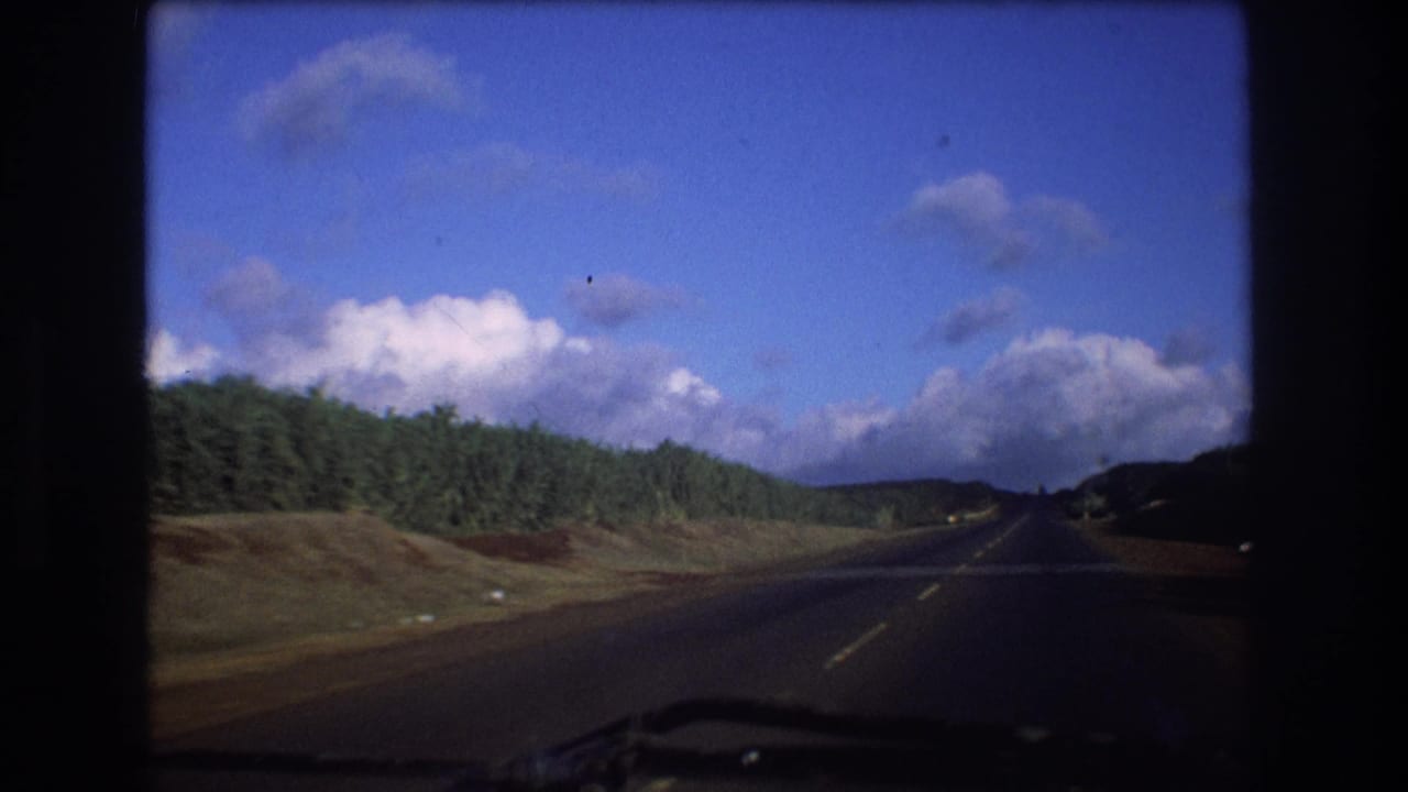 Two-lane road curves through green roadside vegetation beneath scattered clouds.