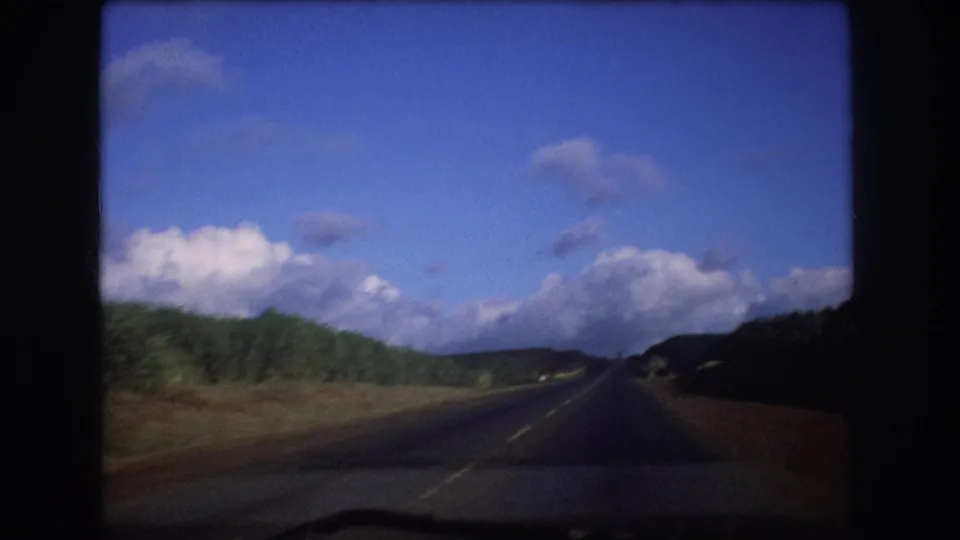 1977 Oahu home-movie frame showing a moving passenger view of a road bordered by green trees and a low hillside under a bright cloudy sky.