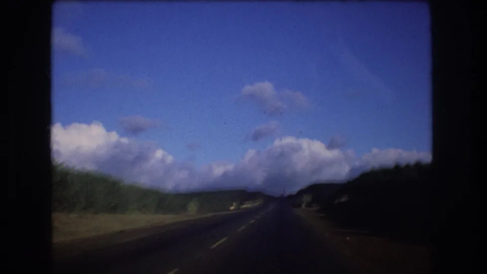 1977 Oahu frame with a slightly darker windshield view down the road toward low hills, roadside vegetation, and large clouds in a blue sky.