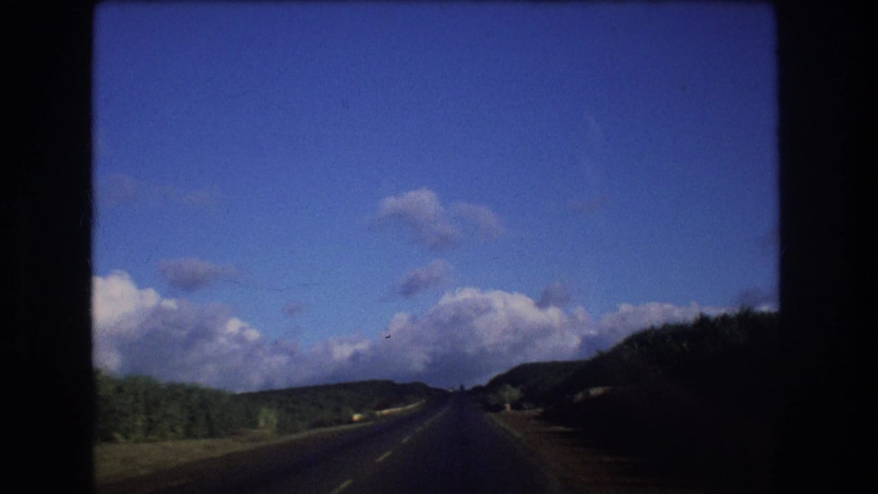 1977 Oahu home-movie frame from inside a moving vehicle, showing a dark-edged view of a two-lane road, green hills, and scattered clouds.