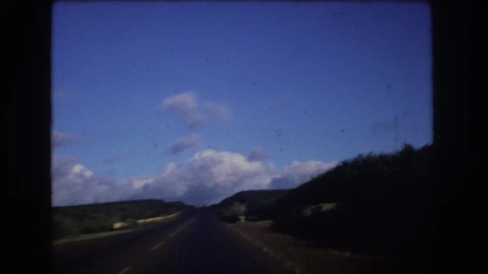 1977 Oahu road-drive view with a paved roadway receding into green hills beneath a bright, clouded sky and heavy 8mm frame borders.