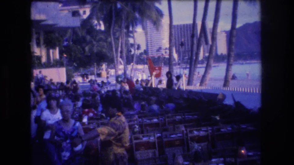 1977 Honolulu Oahu promenade frame showing beachgoers near rows of outdoor seating, tall palms, shoreline water, resort high-rises, and Diamond Head in the distance.