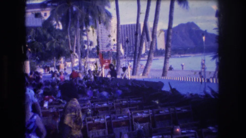 1977 Honolulu Oahu home-movie frame of a crowded palm-lined beachfront promenade, with seating in the foreground, high-rise buildings near the water, and Diamond Head visible beyond.