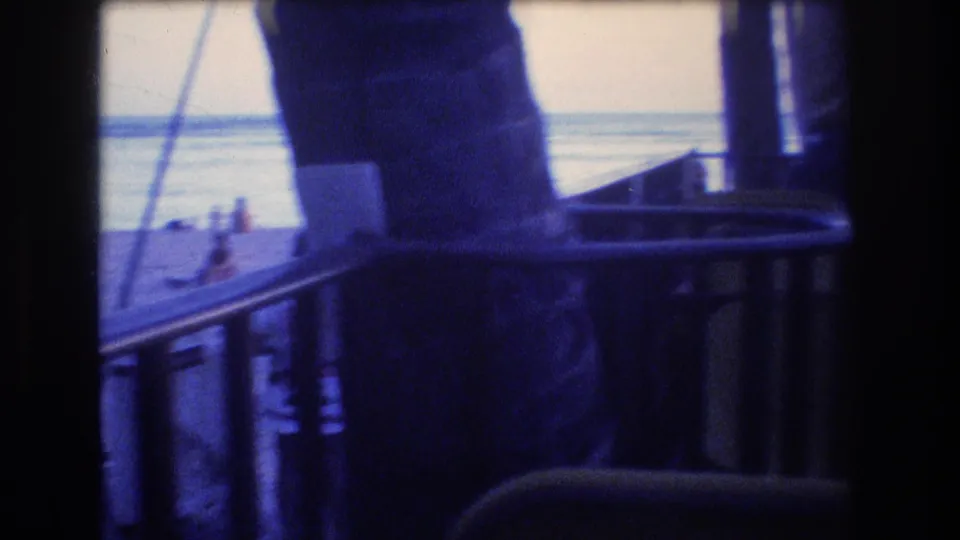 1977 Honolulu 8mm frame showing a darkened boat or pier deck with chair backs, railings, vertical supports, and the ocean horizon beyond.