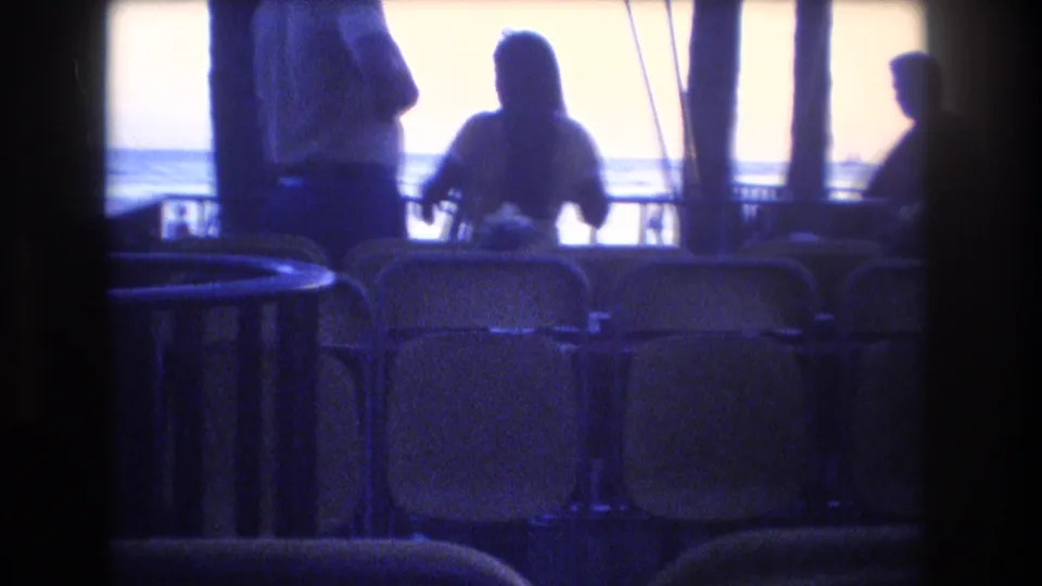 1977 Honolulu home-movie frame with silhouetted figures near rows of yellow chairs, railings, rigging or posts, and the ocean in the background.