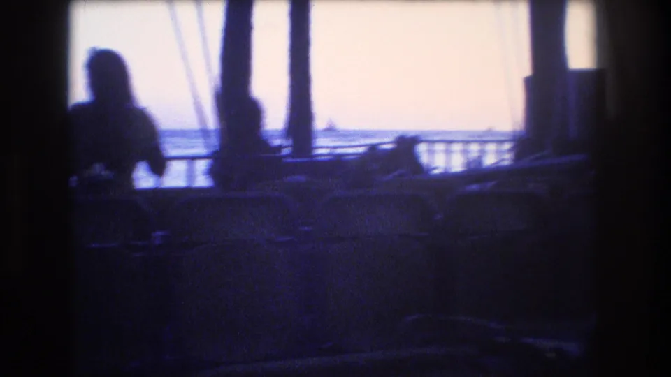 1977 Honolulu 8mm frame showing a silhouetted person near the railing, chair backs in the foreground, vertical rigging, and bright ocean water beyond.