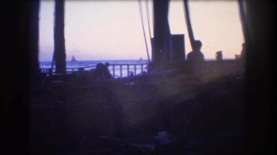 1977 Honolulu home-movie frame looking past dark deck posts, railings, and seated silhouettes toward the ocean horizon in late-afternoon light.