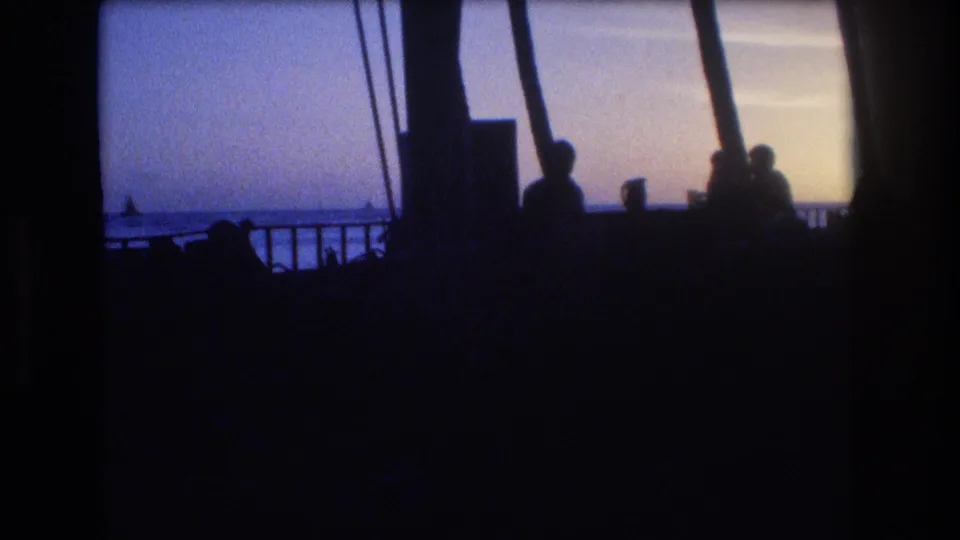 1977 Honolulu Oahu home-movie frame showing dark silhouetted figures by a shoreline railing, with palm trunks and a dim ocean horizon beyond.