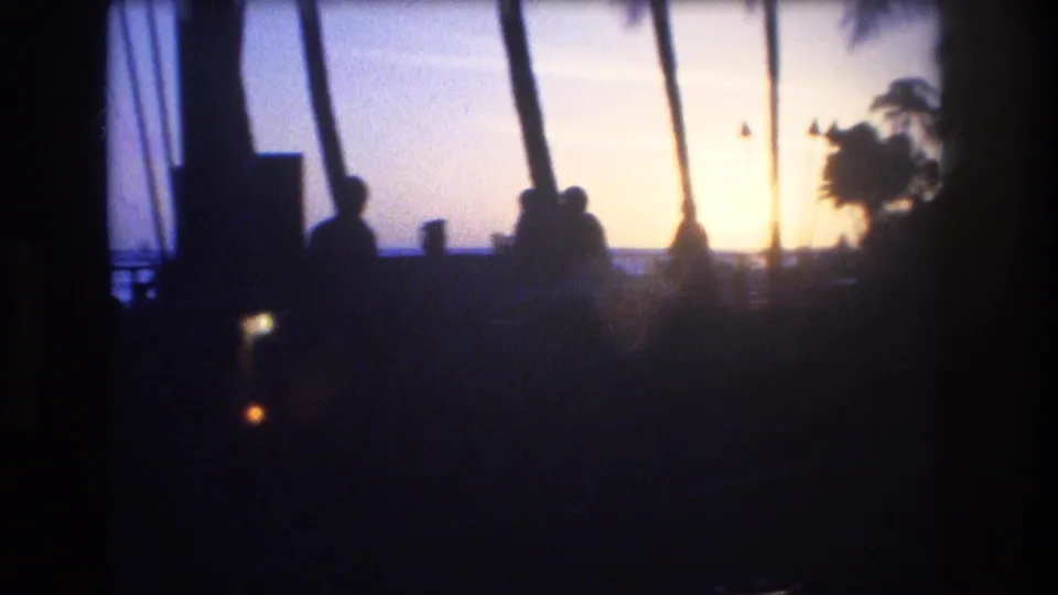 1977 Honolulu Oahu frame with several people in silhouette beneath palms, looking toward the ocean as the sun sets behind the shoreline.