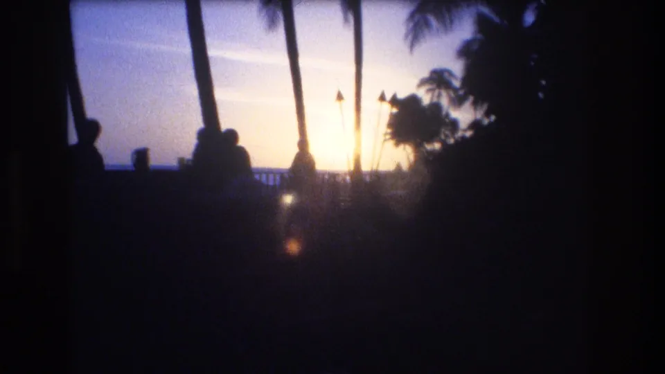1977 Honolulu Oahu home-movie frame showing silhouetted people beside palm trunks, with a bright dusk sky and ocean line beyond the railing.